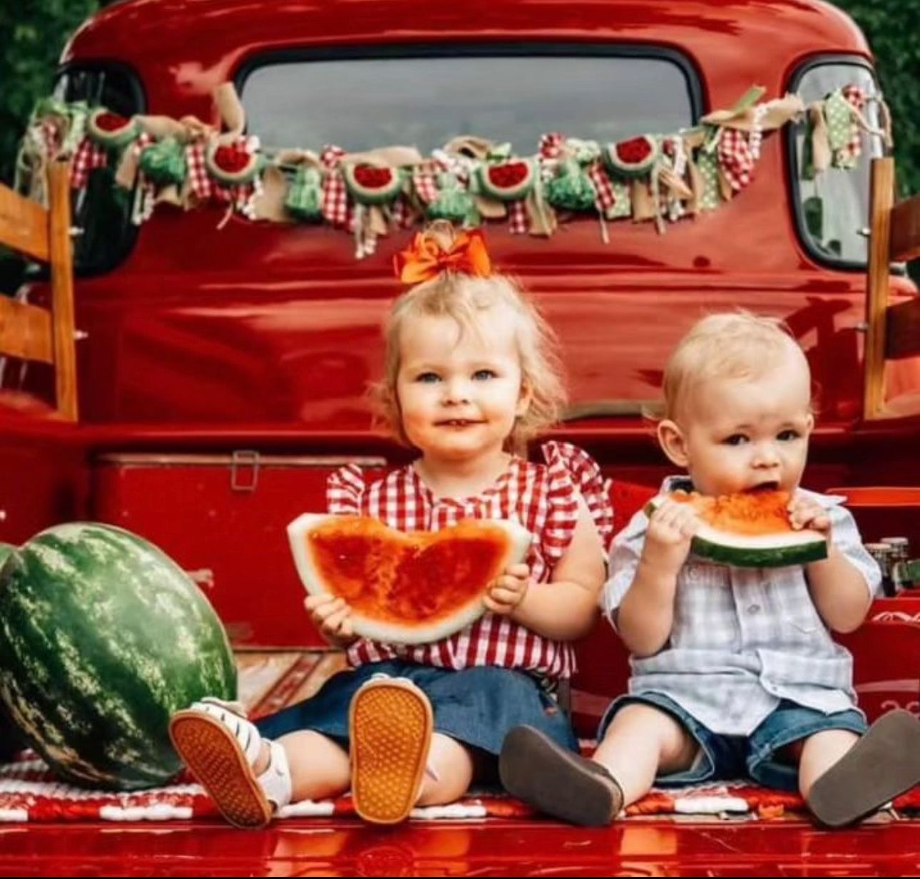 Two toddlers sitting on a red truck eating watermelon slices outdoors, with a festive watermelon garland backdrop.