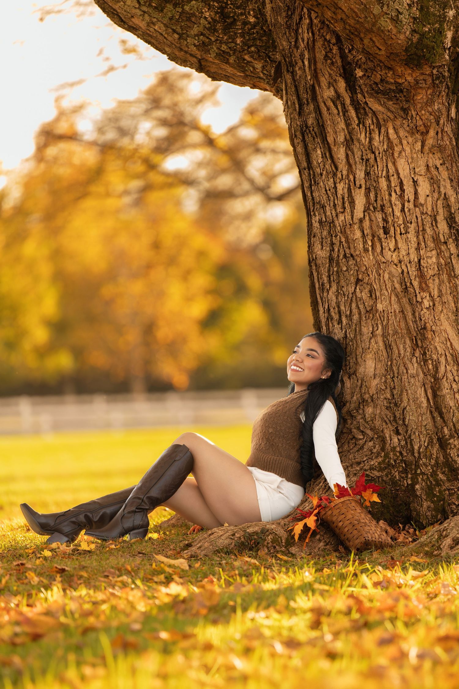 Woman in autumn park leaning against large tree trunk, wearing boots and brown vest, smiling among fall foliage.