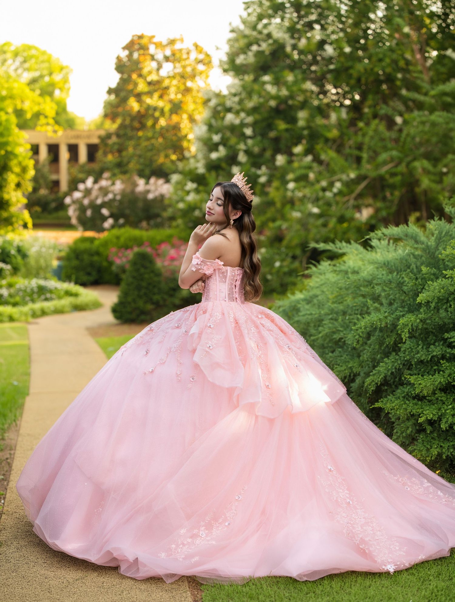 Young woman in a pink ball gown and tiara poses gracefully in a lush garden for a quinceañera photo.
