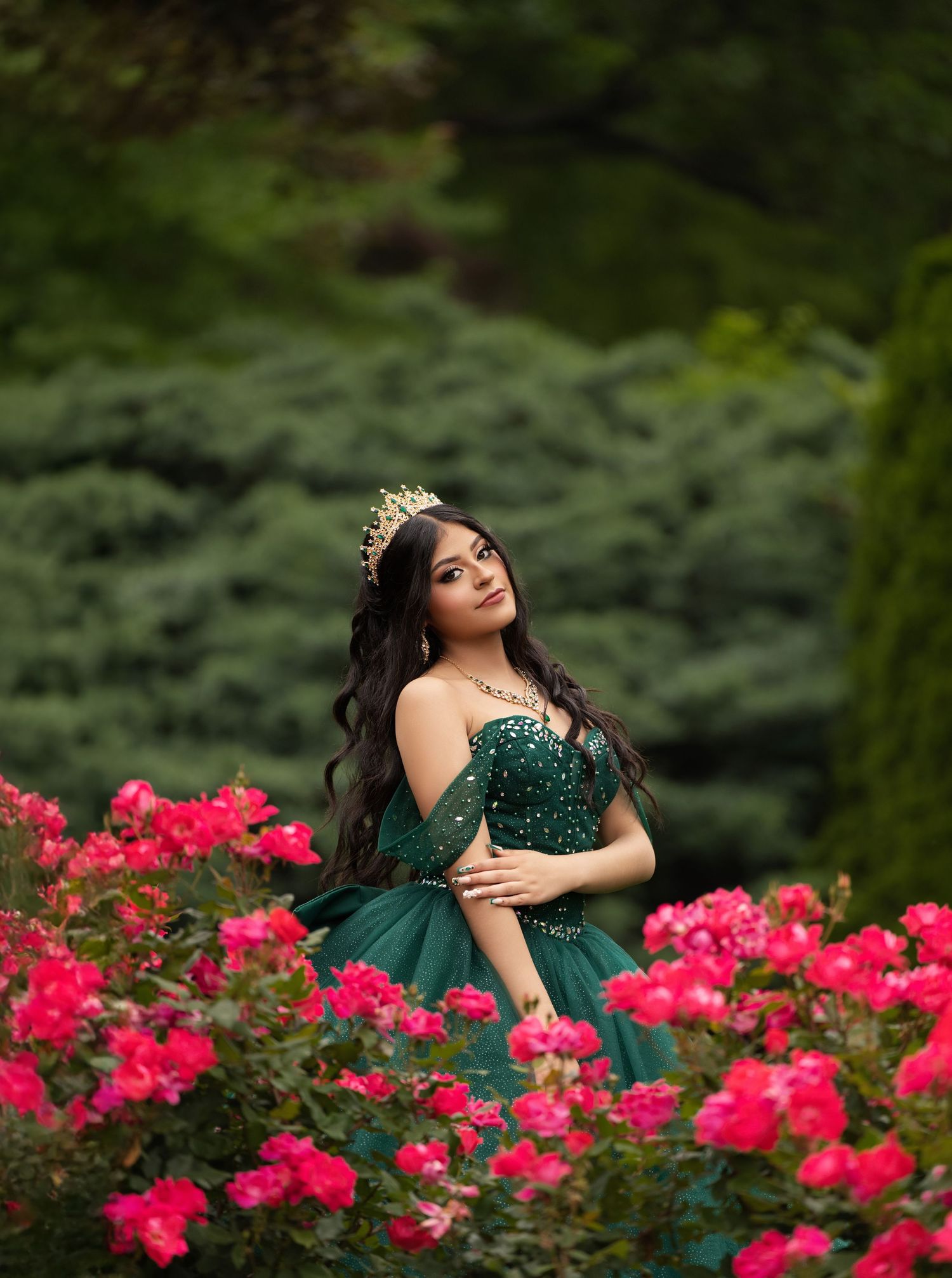 Young woman in emerald green ball gown and tiara poses gracefully among vibrant pink roses in a lush garden.