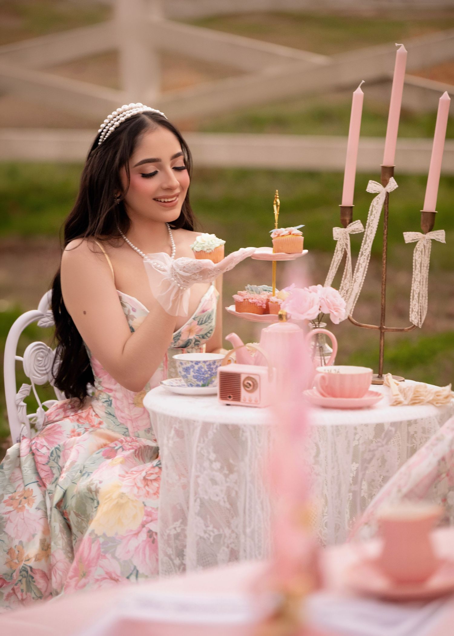 Young woman in floral dress holding cupcakes at elegant pink tea party table with candles and tiered dessert stand.
