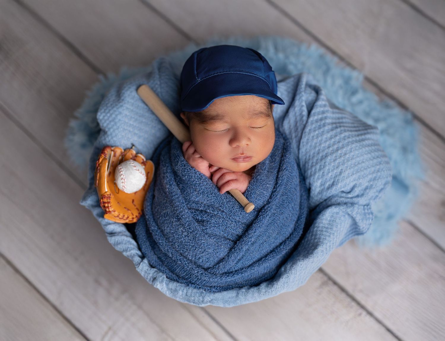 Baseball themed newborn photo with blue wrap, cap, and miniature glove on wooden floor.