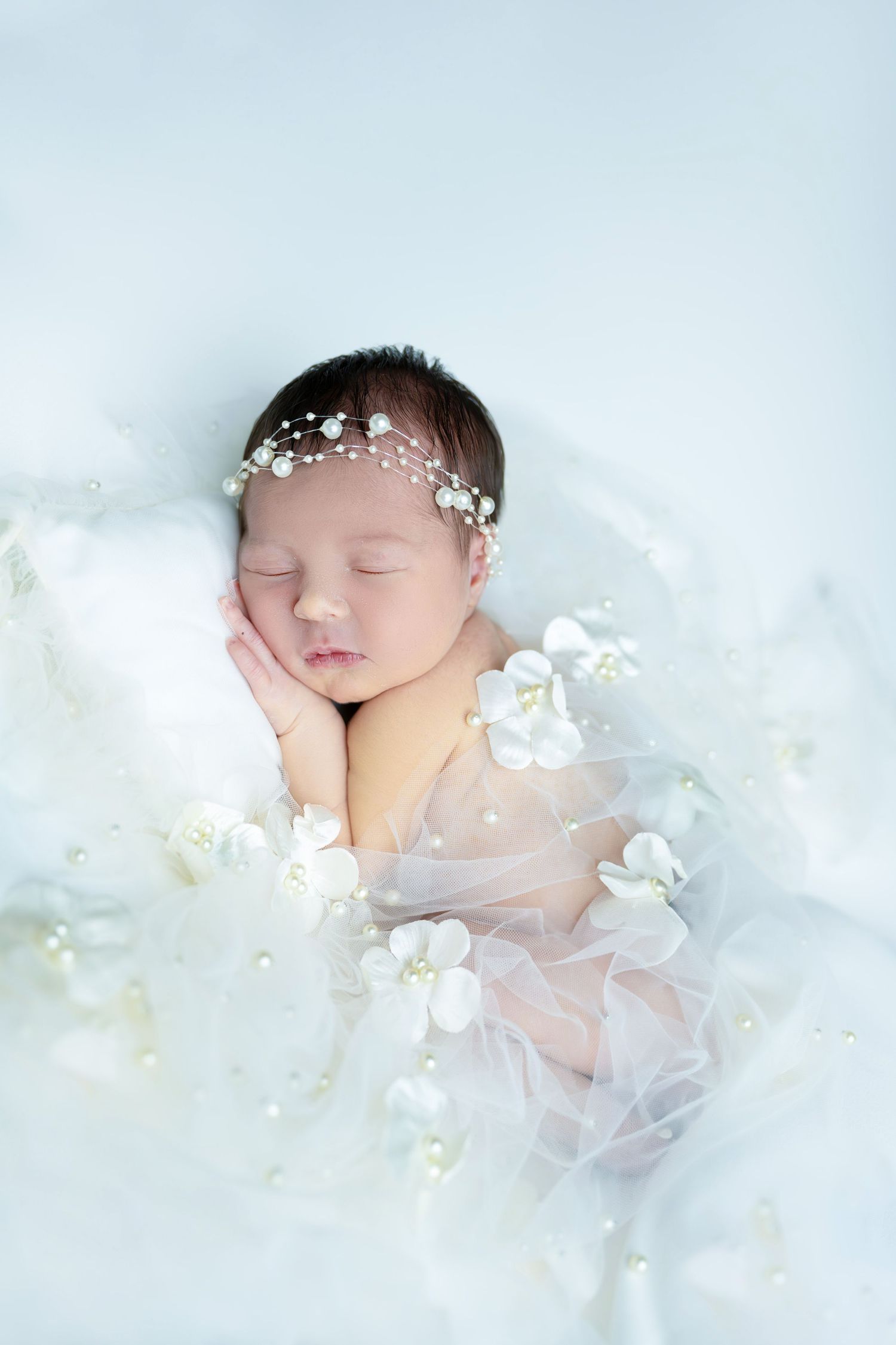 Sleeping newborn with pearl headpiece surrounded by white orchids and dreamy tulle.