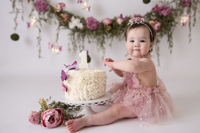 A baby in a pink tutu dress poses with floral decorations and a birthday cake in a light studio setting.