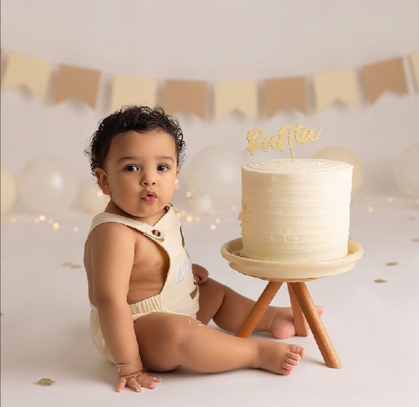 Minimalist white cake smash photo setup with wooden cake stand and gold banner decorations in the background.