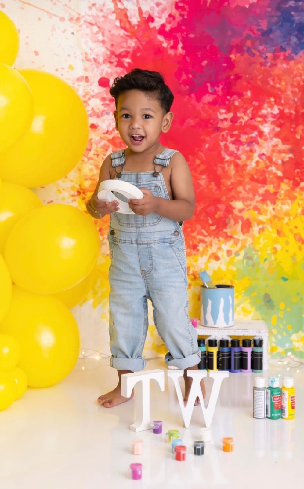 Colorful paint splatters create vibrant backdrop for creative photo session with small child in denim overalls.