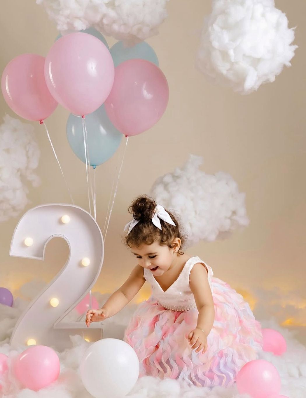 A light-up number 2 with pink and blue balloons, white pom poms, and a child in a tulle dress for birthday photos.