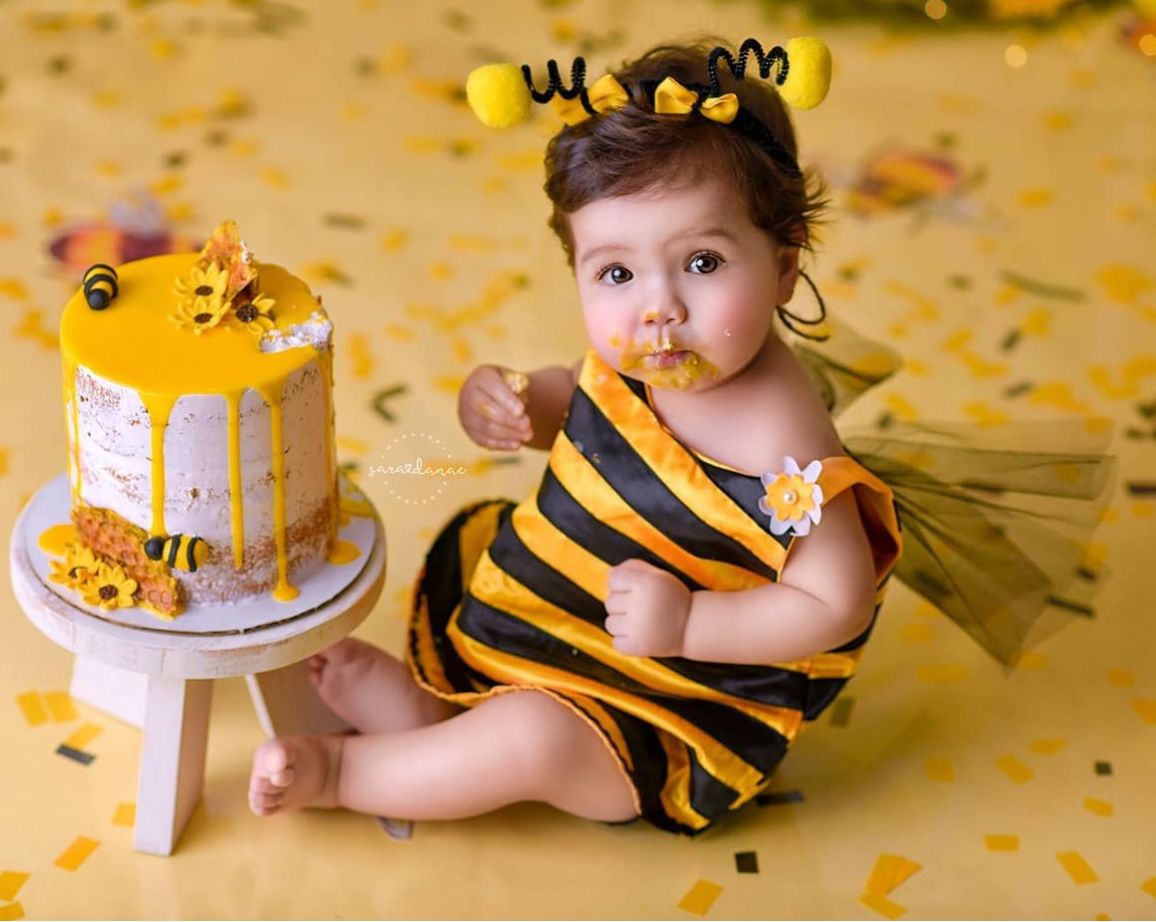 A baby wearing a yellow and black striped bee costume sits on the floor next to a birthday cake with confetti scattered around.