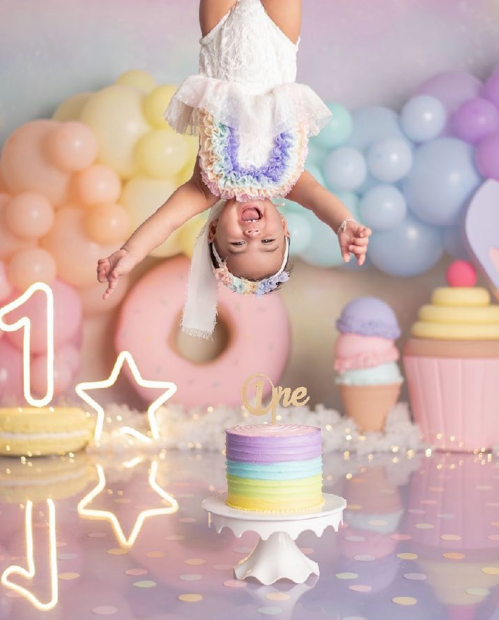 Whimsical first birthday party setup with pastel balloons, rainbow cake, and upside-down pose against a dreamy backdrop.