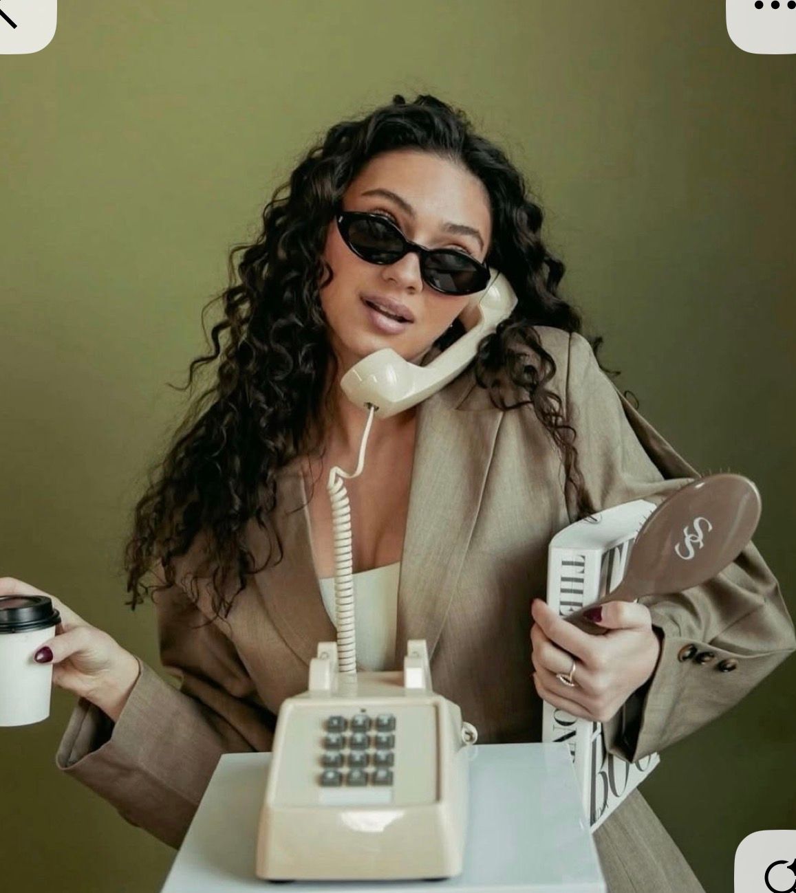 Woman in sunglasses and blazer poses with vintage phone, coffee cup, and mirror in a stylish retro-themed photo shoot.