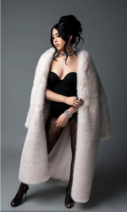 Woman in a black bodysuit, lace tights and heels poses in a long white faux fur coat with a sparkling choker against a gray studio backdrop.