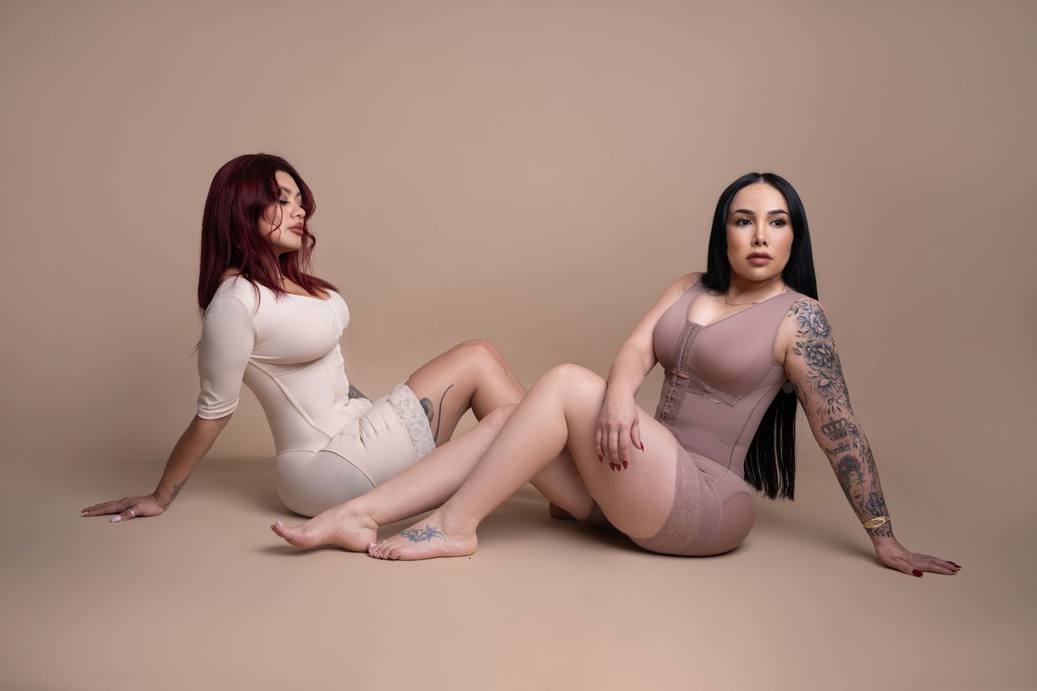 Two women in matching nude bodycon outfits sit on a beige studio floor, one with red hair and one with long black hair and arm tattoos.