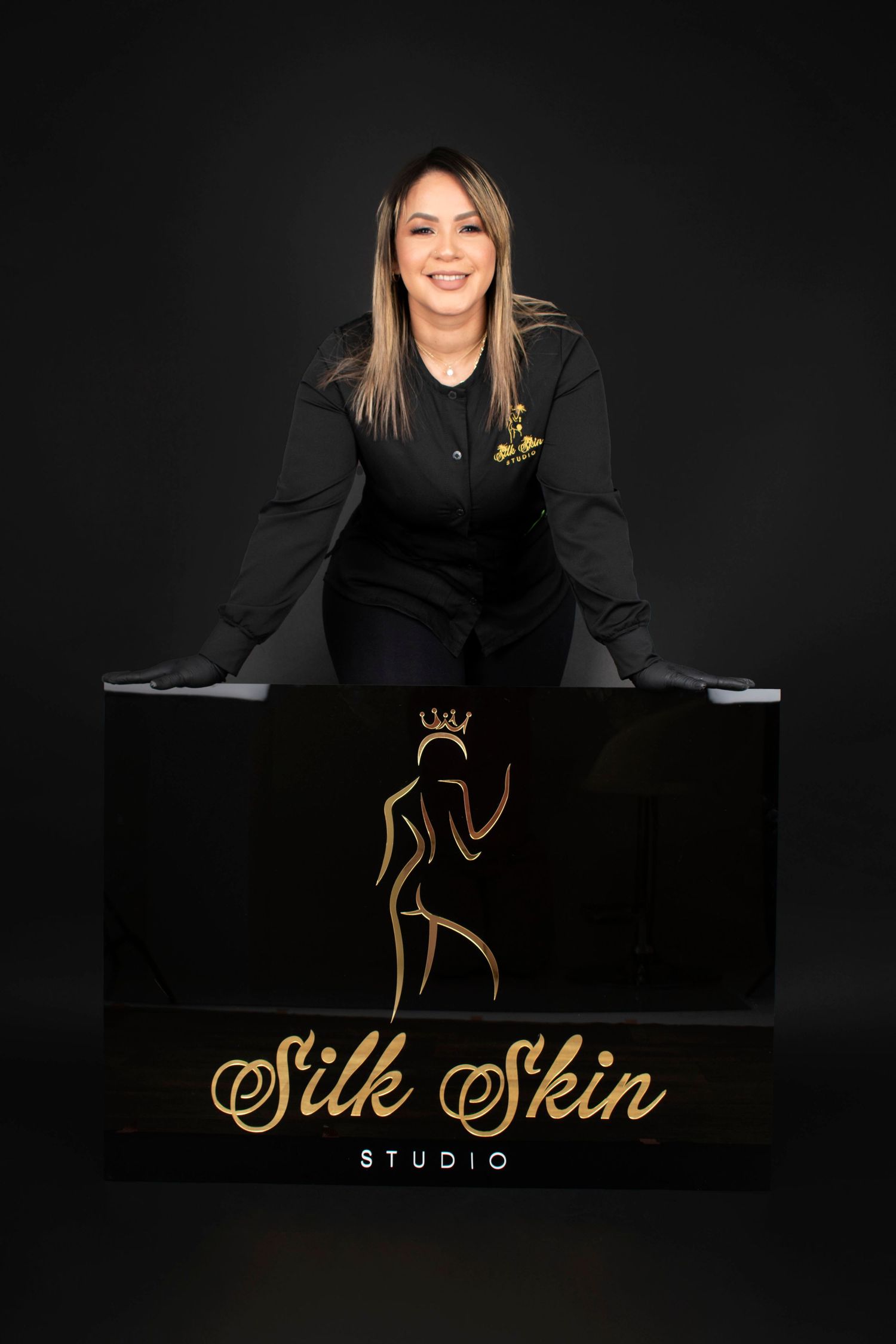 Smiling esthetician in a black uniform leans on a black and gold Silk Skin Studio sign with a crown logo against a dark background.
