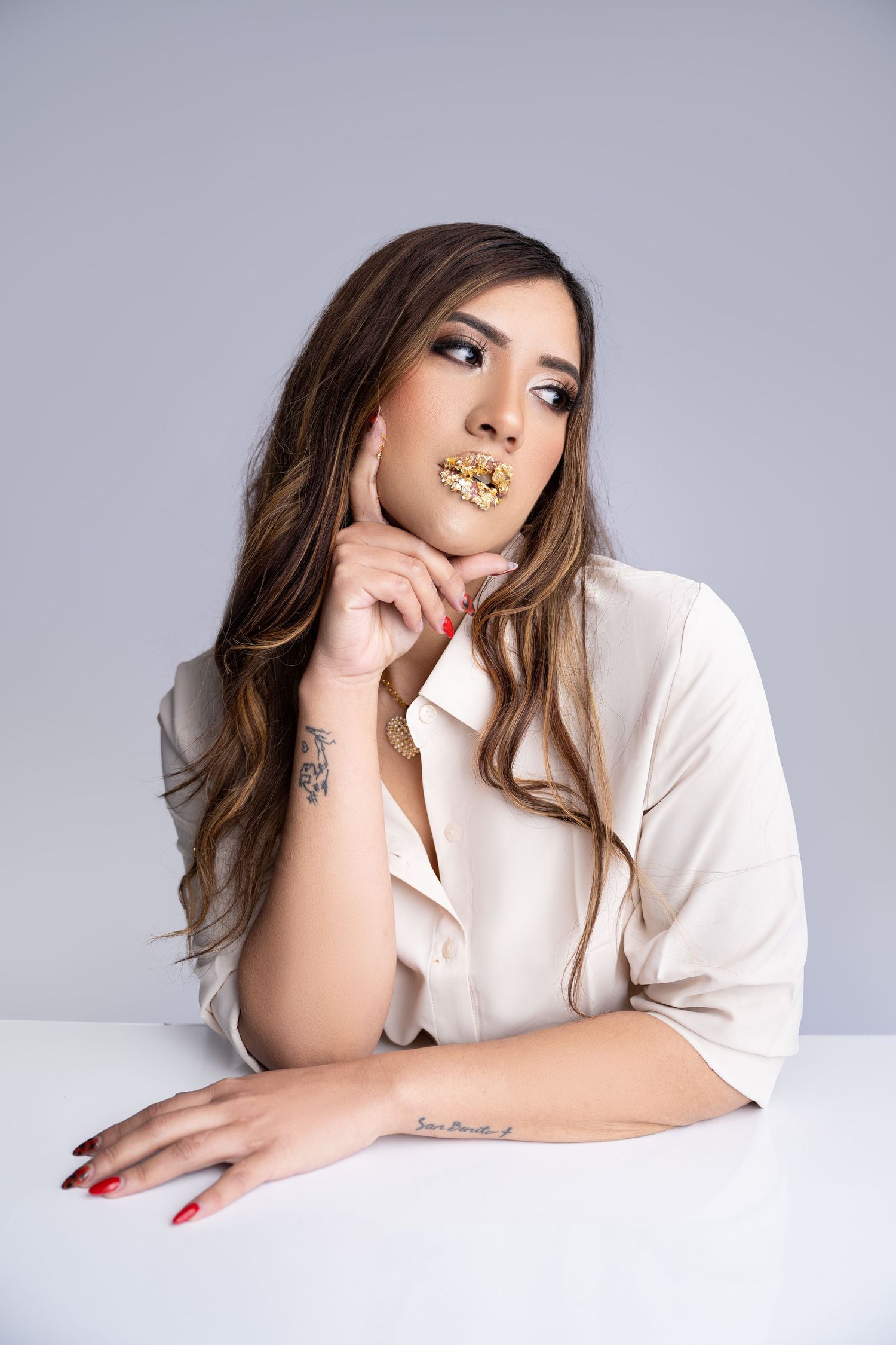 Young woman with gold lip makeup and tattoos sits at white table in beige blouse, posing thoughtfully against gray background.