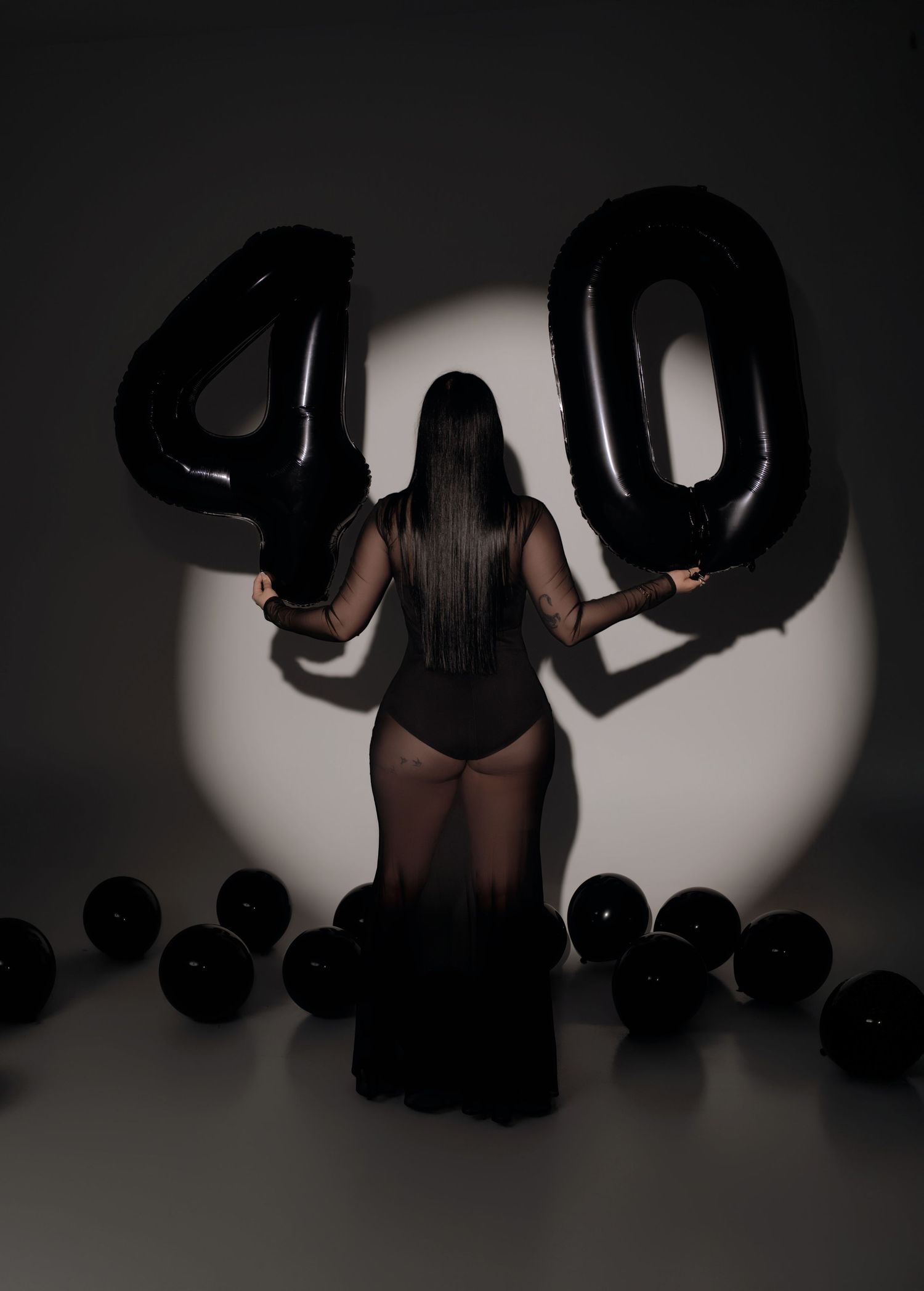 Woman holding black number 40 balloons from behind, surrounded by black balloons in dramatic spotlight for a birthday celebration.