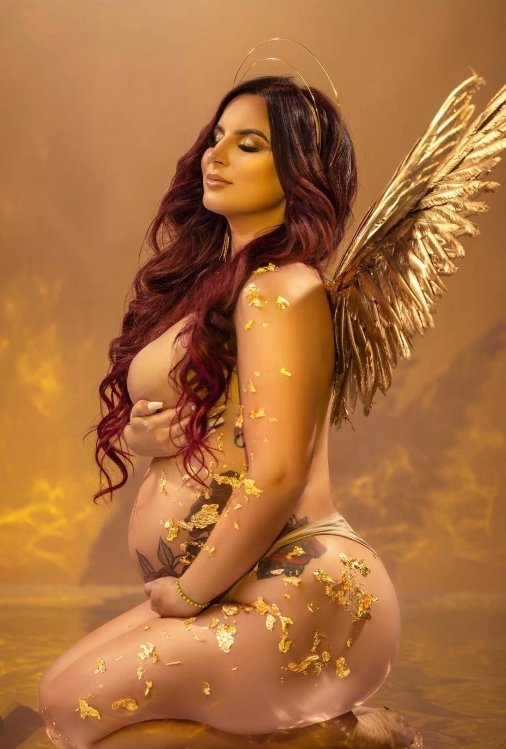 Woman with red hair poses as golden angel with wings and halo, adorned with gold leaf flakes in artistic maternity photo.