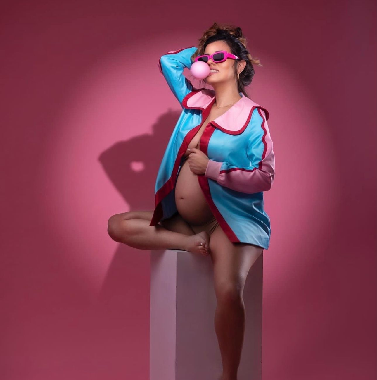 Pregnant woman in blue pink robe with sunglasses poses on white pedestal against pink background.