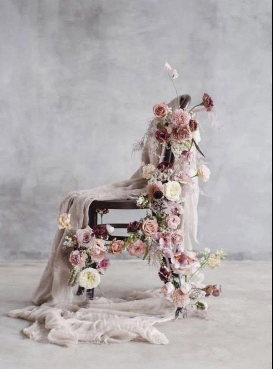 A wooden chair draped with blush fabric and adorned with cascading pink, mauve, and cream roses against a gray backdrop.