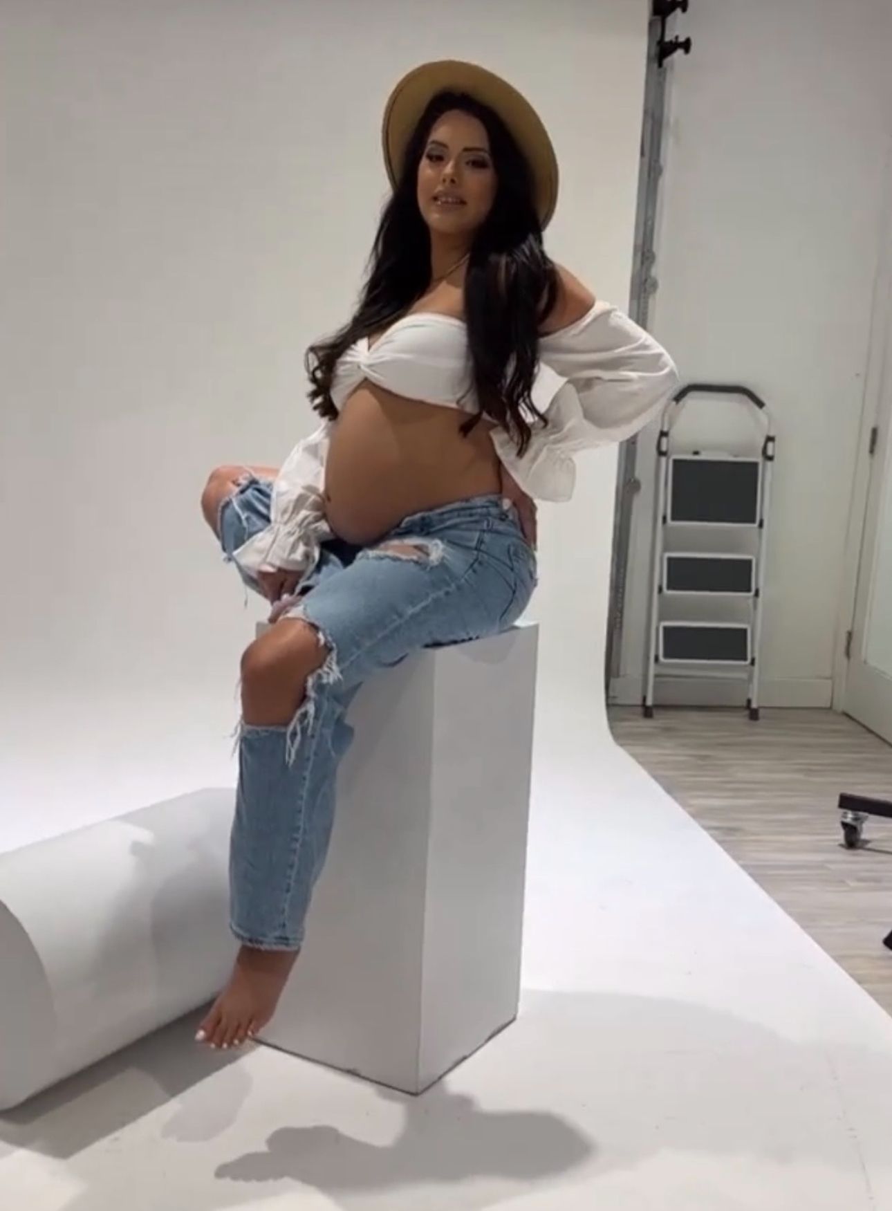 Pregnant woman in white crop top and distressed jeans sitting on white cube in photo studio.