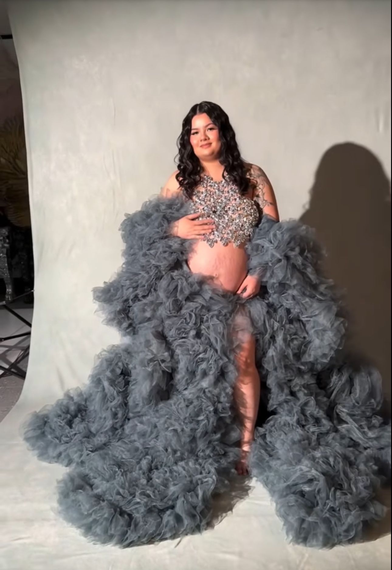 Pregnant woman posing in a stunning gray ruffled tulle maternity gown with beaded bodice in a photo studio.