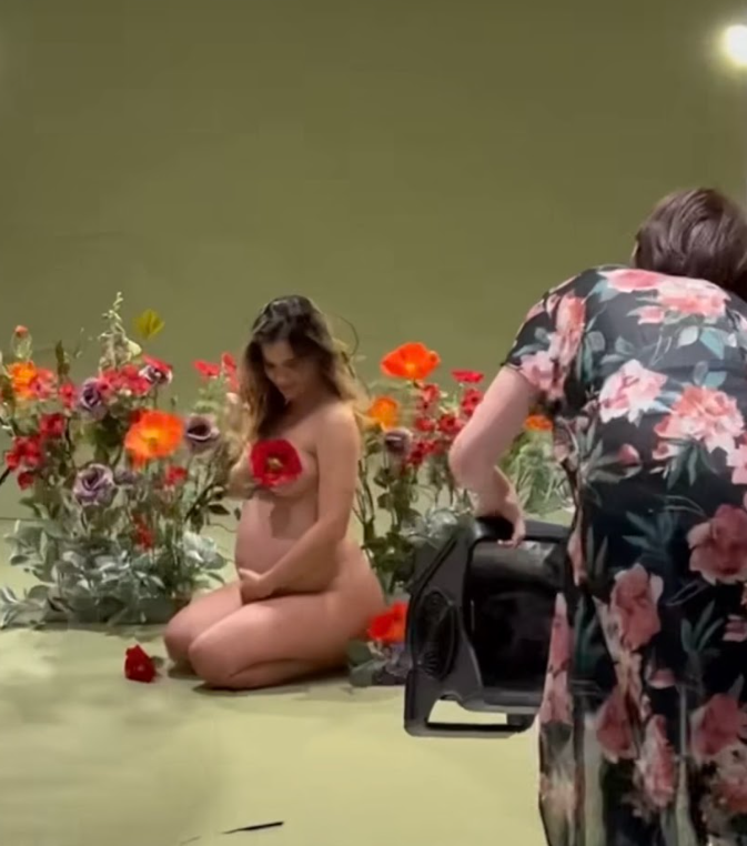 A person in a floral dress lies on the ground near colorful potted flowers in a garden setting.