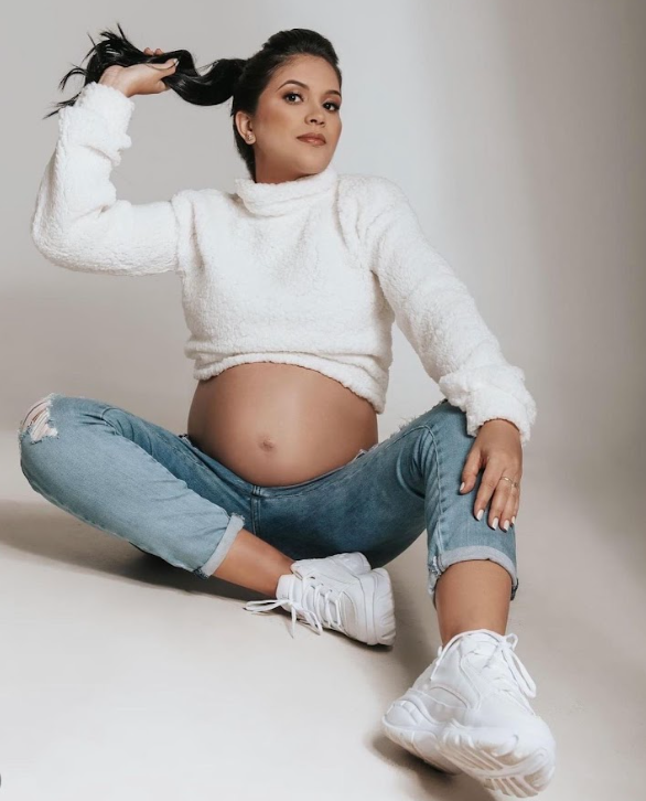 Maternity photo series showing expectant mother in white sweater and jeans sitting on floor against neutral backdrop.