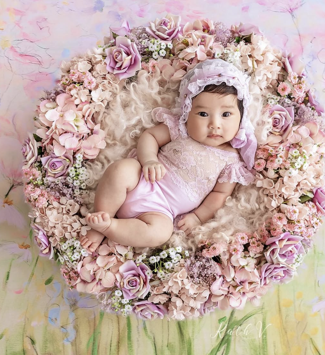 Adorable baby in pink lace bonnet and romper nestled in a round floral wreath of roses.