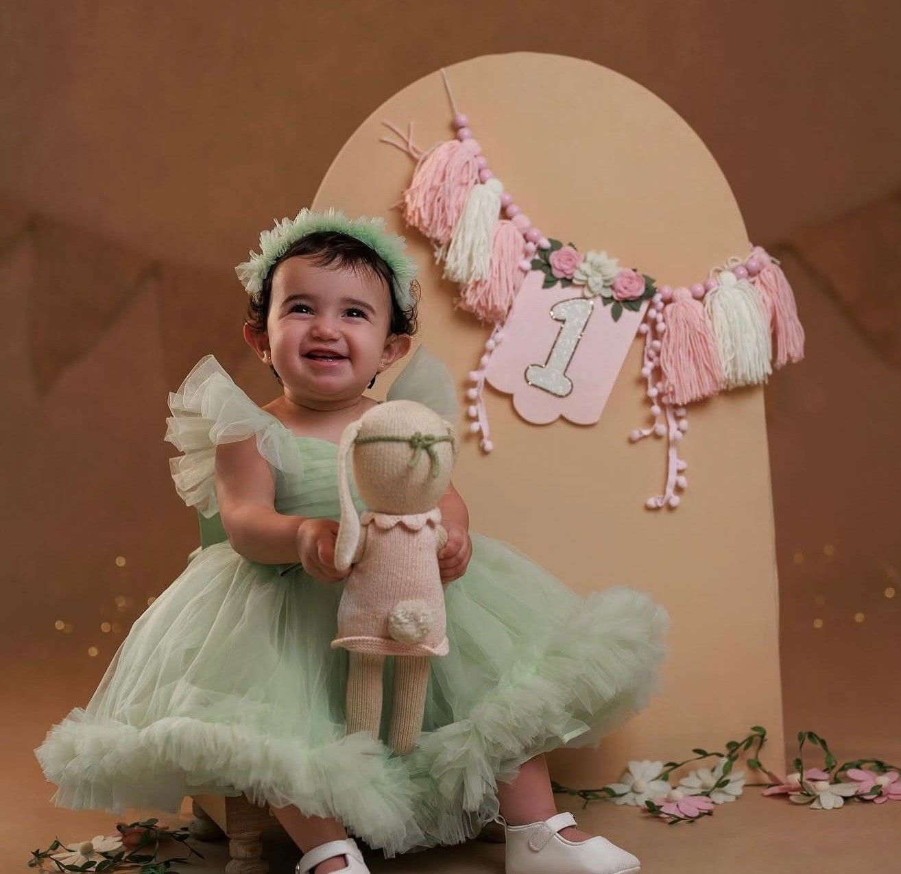 Smiling baby girl in mint green tutu dress holding a doll at her first birthday party celebration.