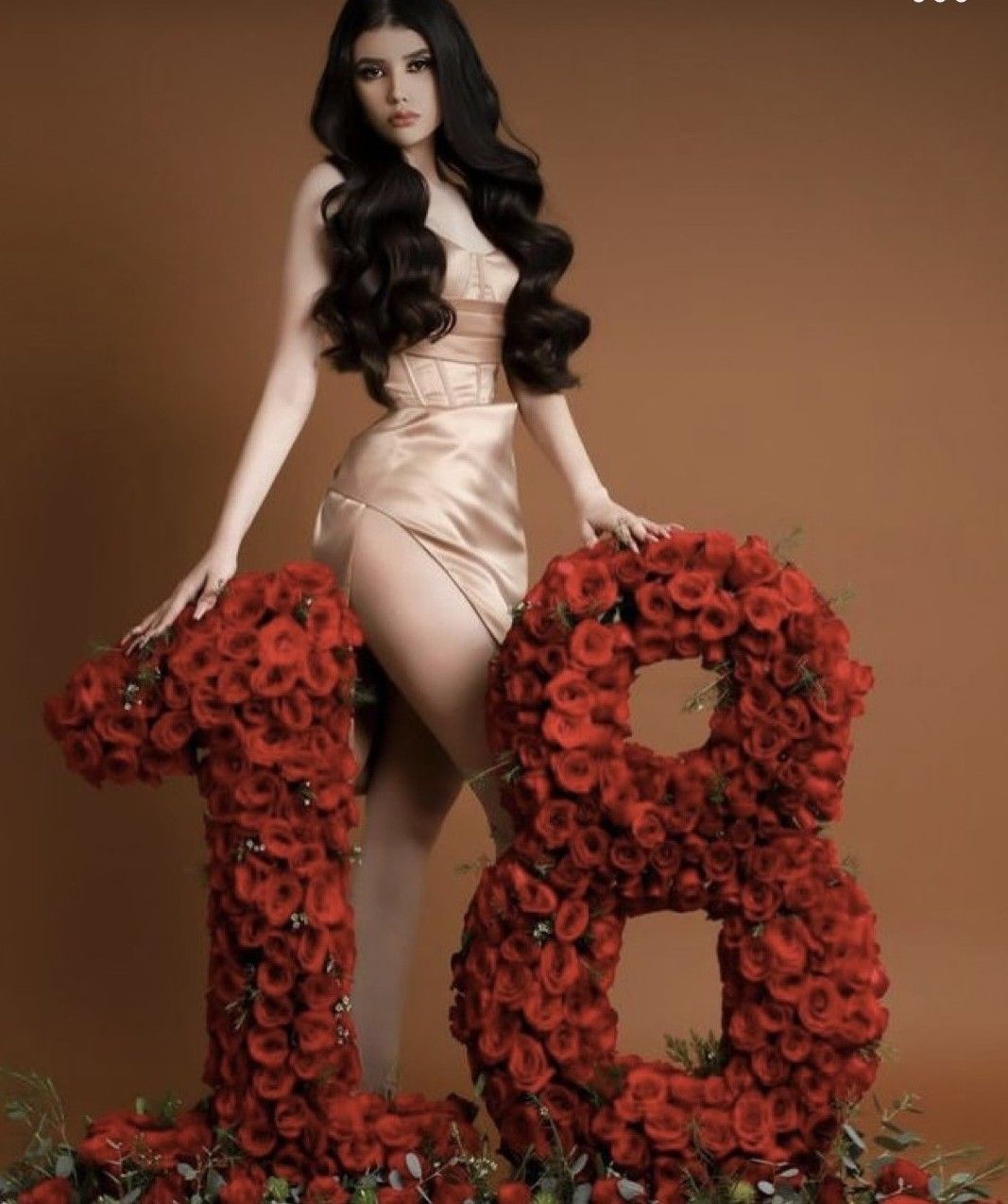 Woman in champagne gown poses with large floral number 18 made of red roses for an elegant birthday celebration.