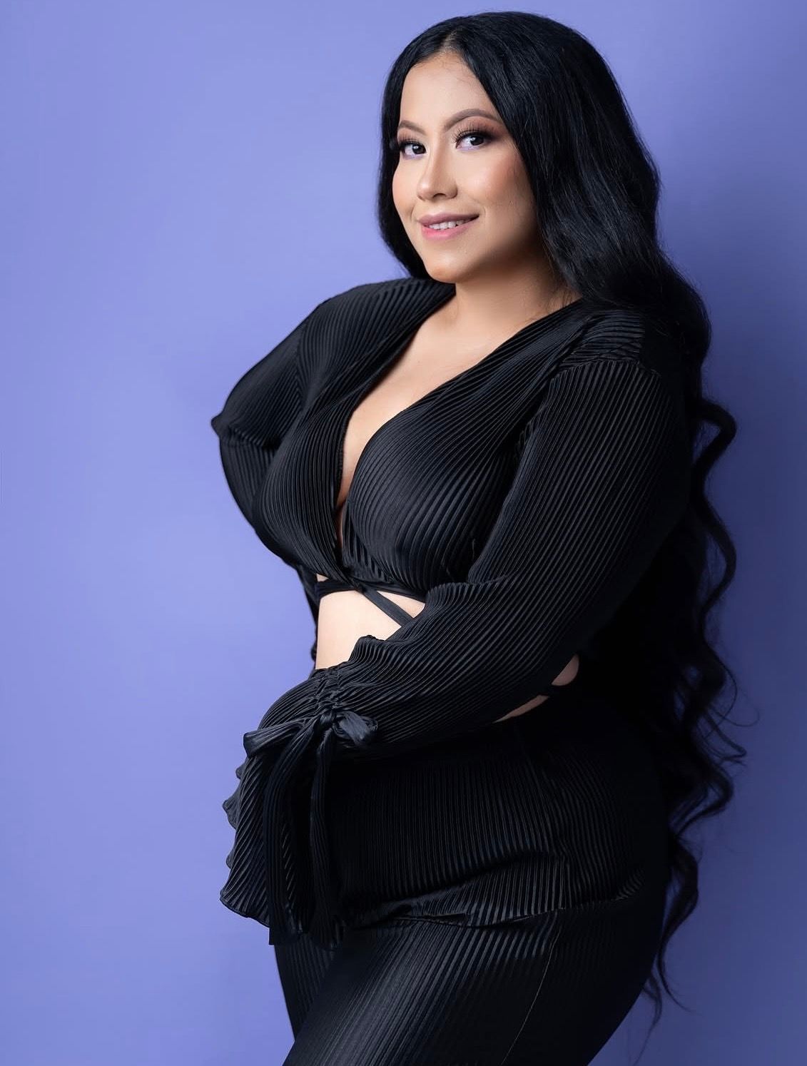 Woman in black plunging neckline ribbed crop top and skirt posing against purple background with long wavy hair.