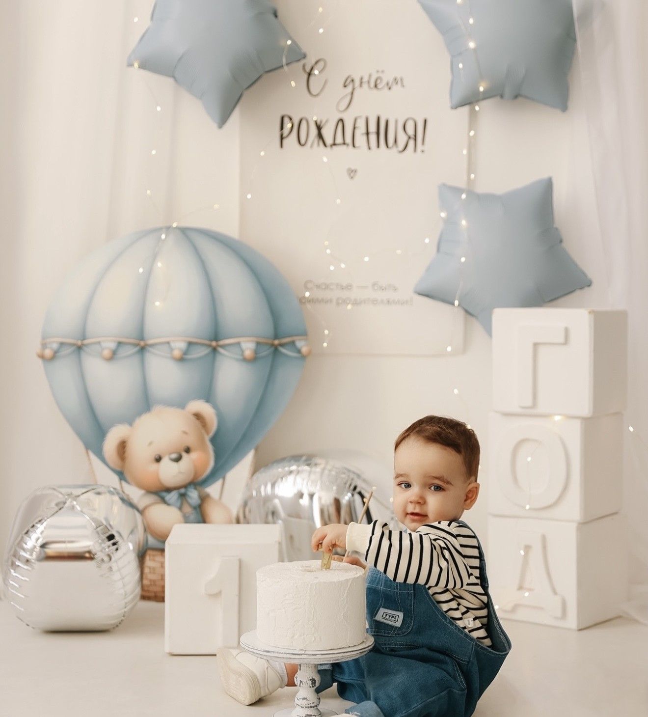 Baby boy celebrating first birthday with white cake, teddy bear balloon, star balloons, and Russian birthday decorations.