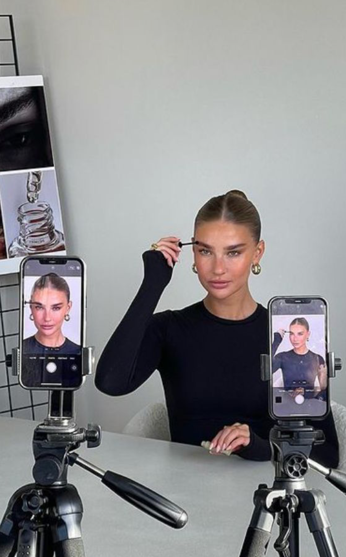 Woman applying makeup while recording herself on two smartphones mounted on tripods at a white desk.