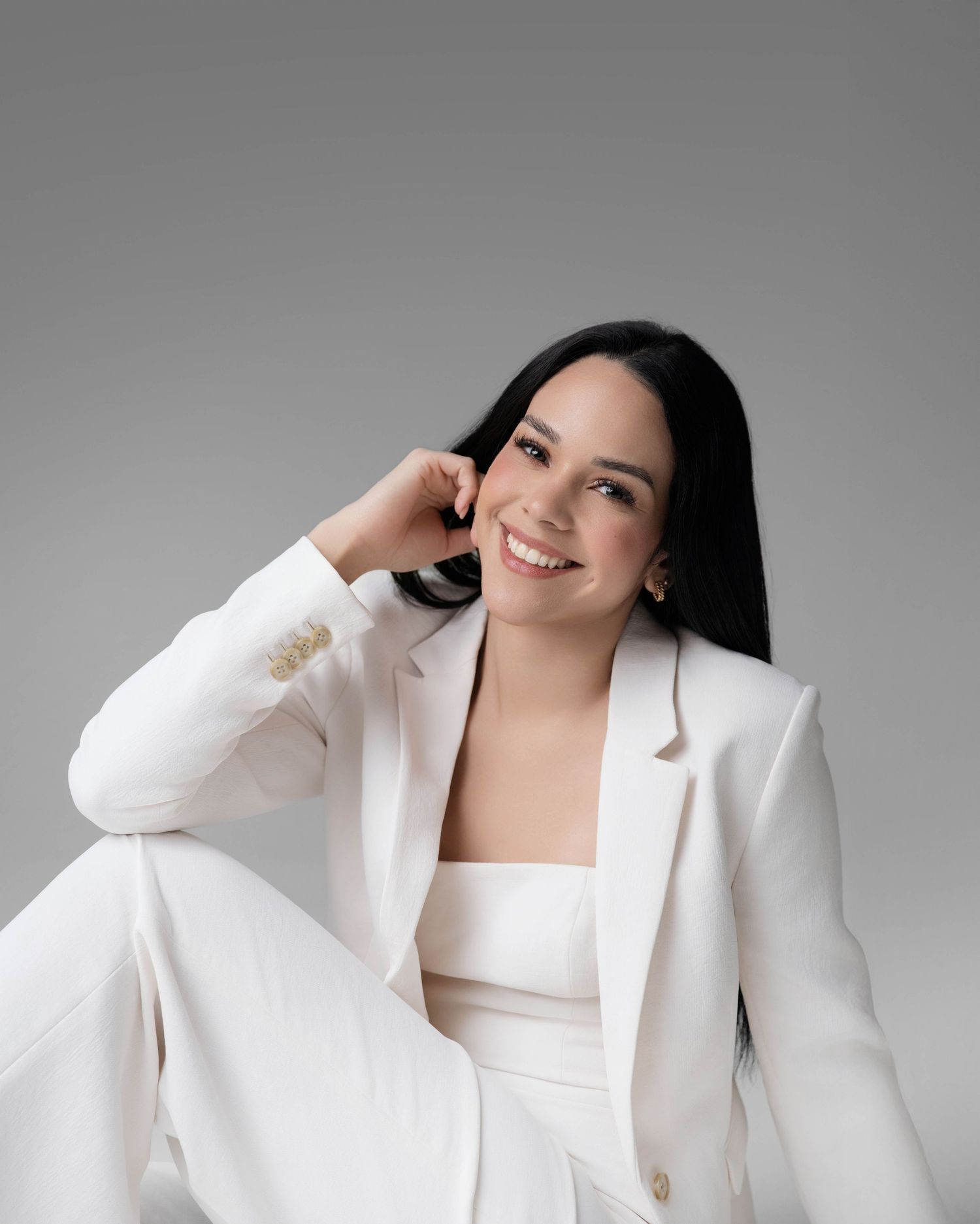 Woman in white blazer suit smiling confidently, seated against gray background in elegant professional attire.