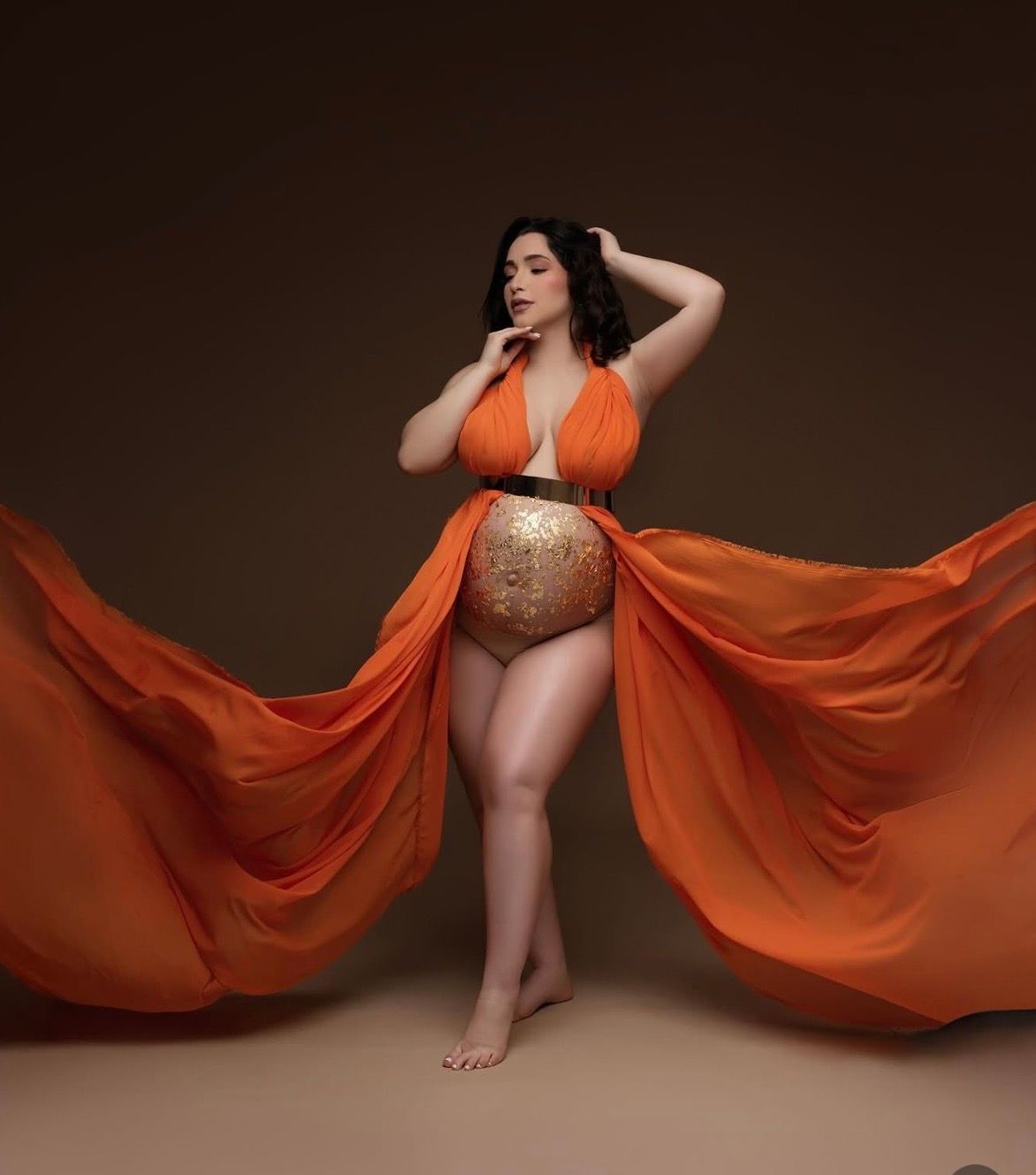 Woman in an orange halter top and flowing orange chiffon skirt with gold sequin details poses barefoot in a studio maternity photoshoot.