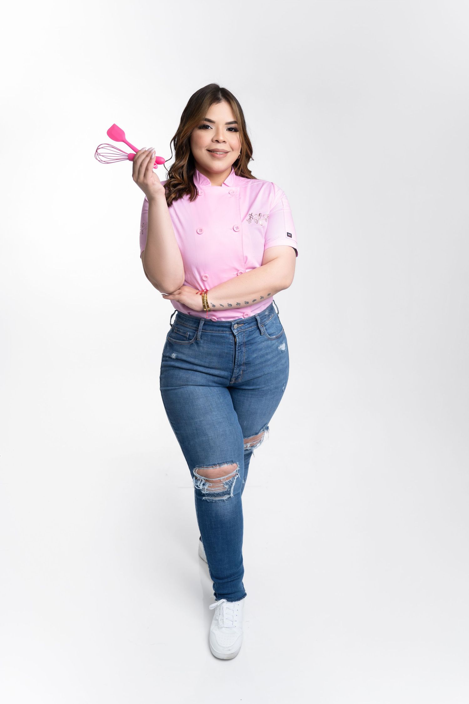 Smiling woman in a pink chef coat, ripped blue jeans and white sneakers holds pink whisk and spatula against a white studio background.