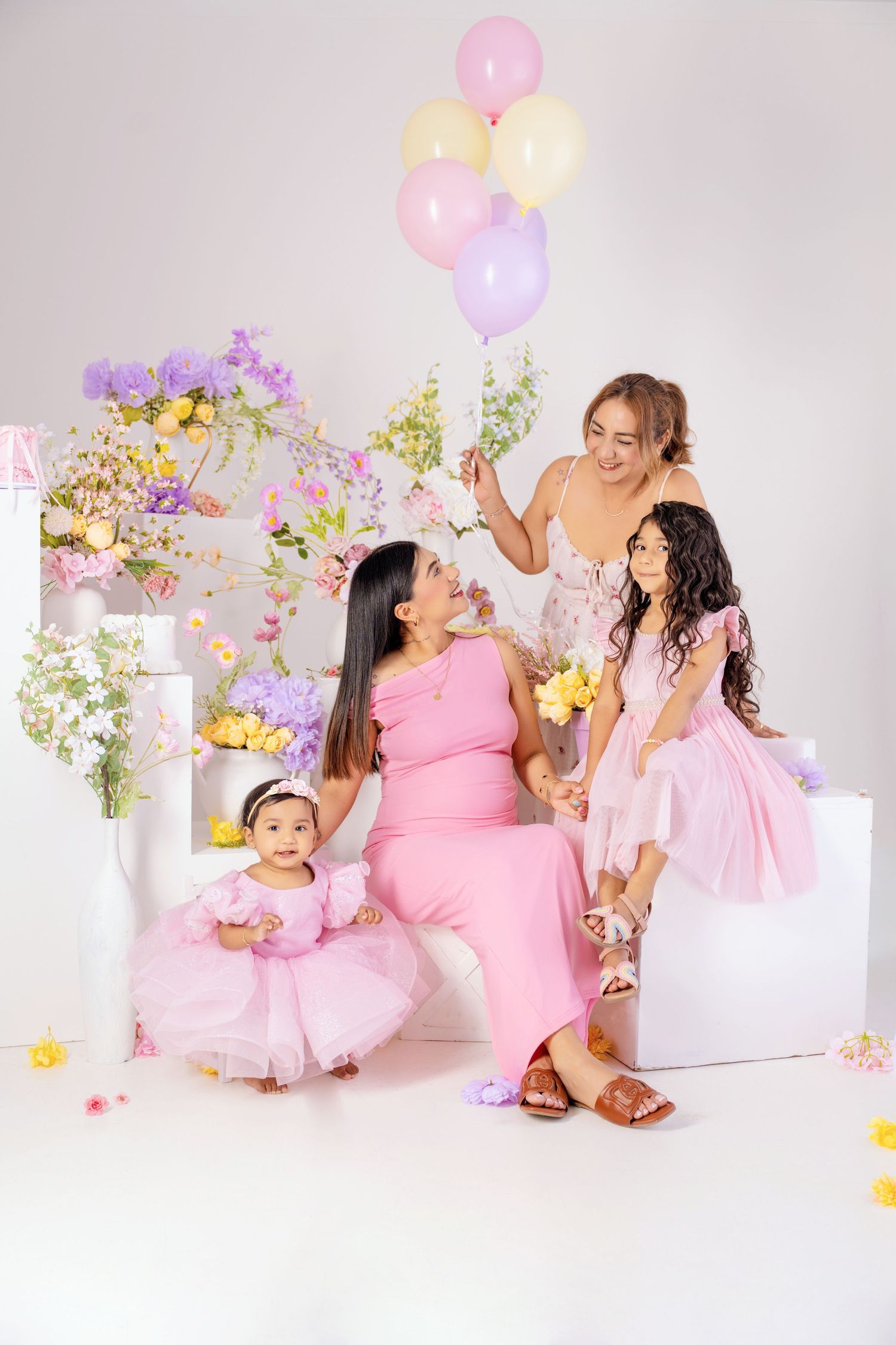 Four women and girls in pink dresses pose with colorful balloons and flowers in a bright studio setting.