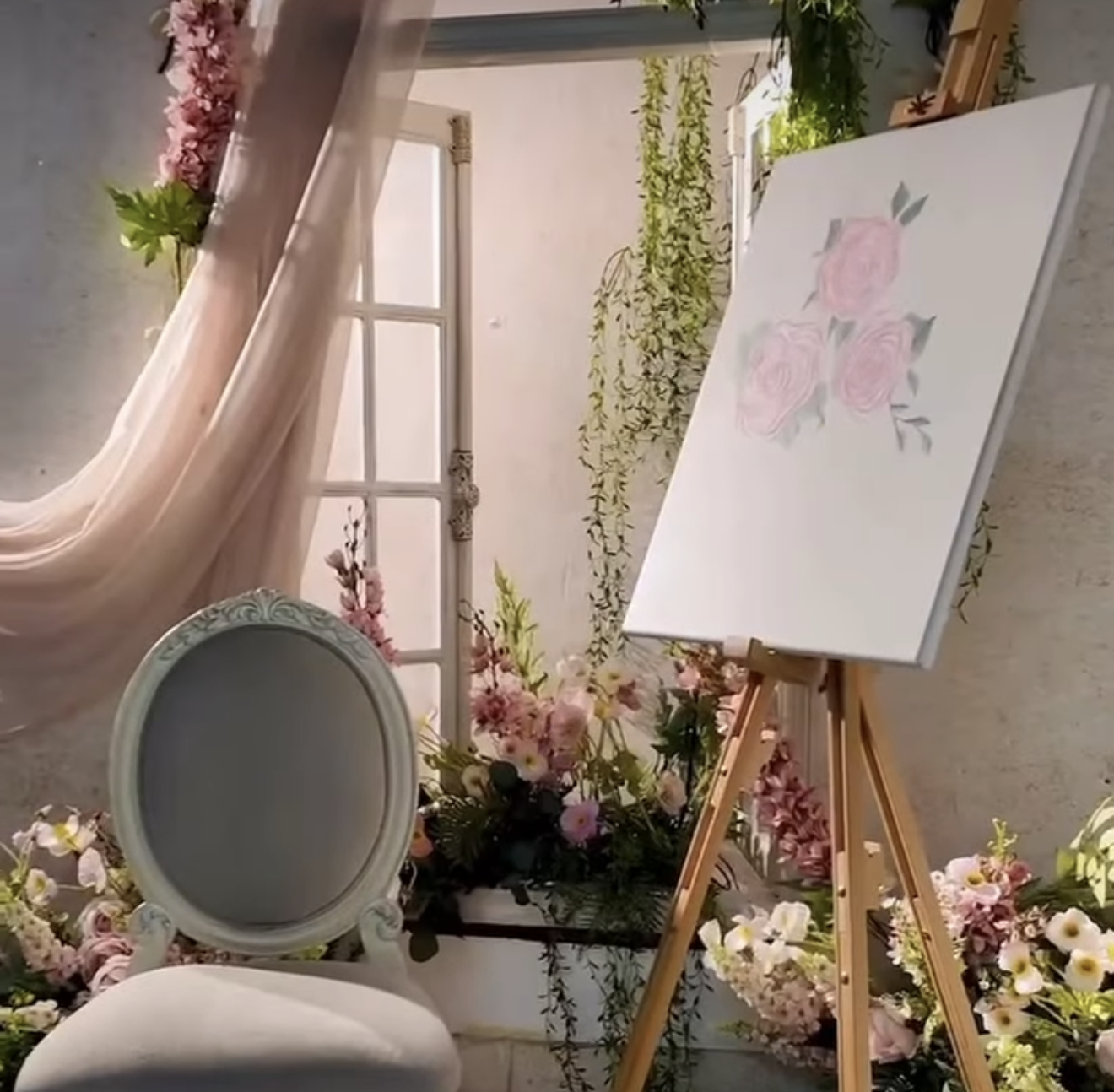 Elegant easel with pink rose painting beside oval mirror, surrounded by lush floral arrangements in a romantic setting.