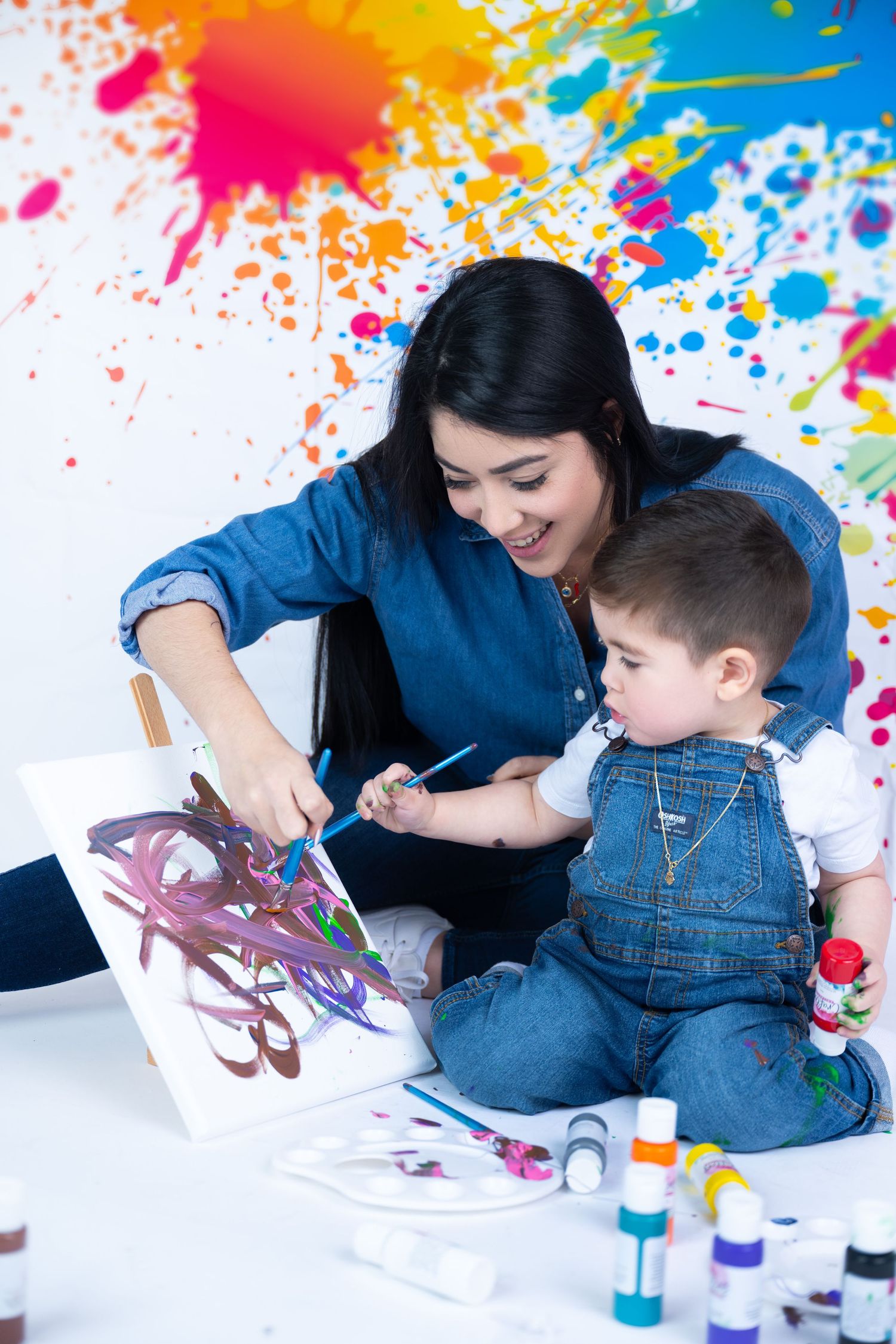 Mother and toddler painting together on canvas with colorful paint splashes on white wall background.