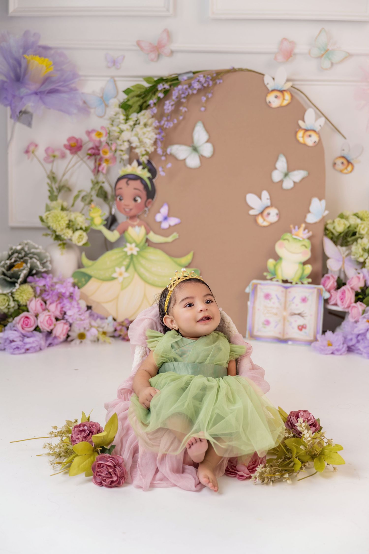 Baby girl in green princess dress sits among flowers with butterfly decorations and Princess Tiana backdrop.