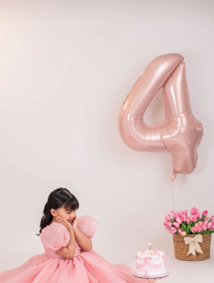 Birthday celebration setup with pink number 4 balloon, pink dress, cake and floral decorations against white wall.