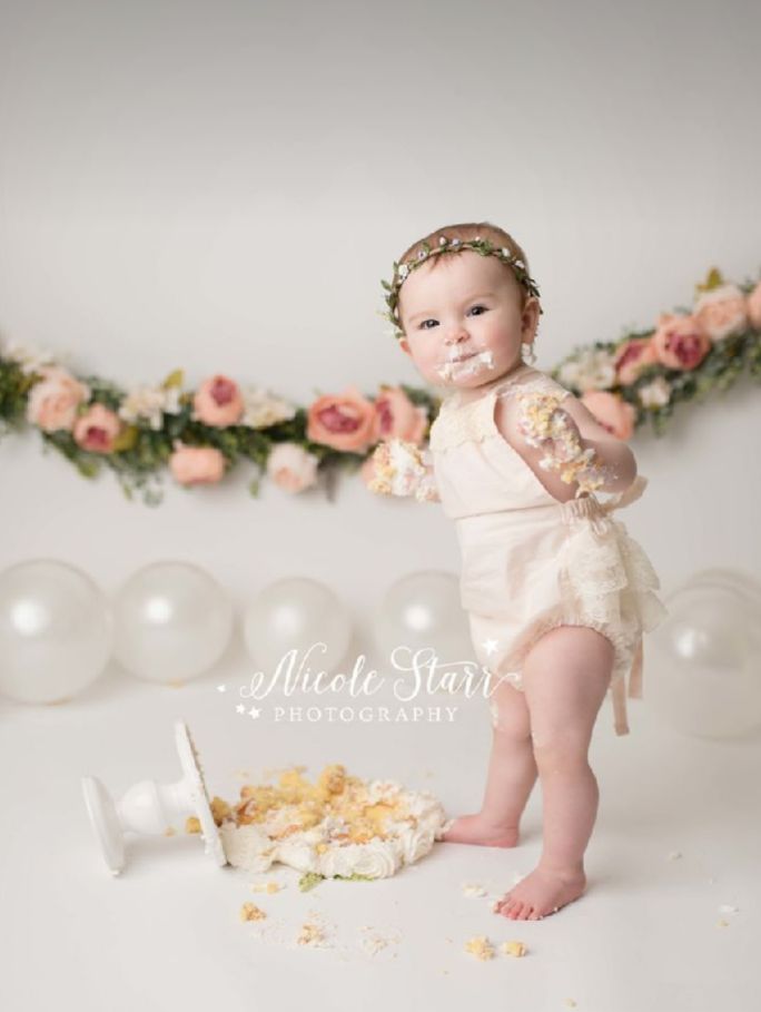 Baby cake smash photography session with pink floral decorations and string lights in the background.