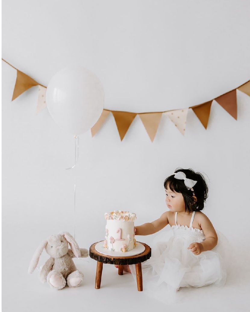 A minimalist first birthday party setup with white bunting, stuffed animals, and a small cake on wooden stool.
