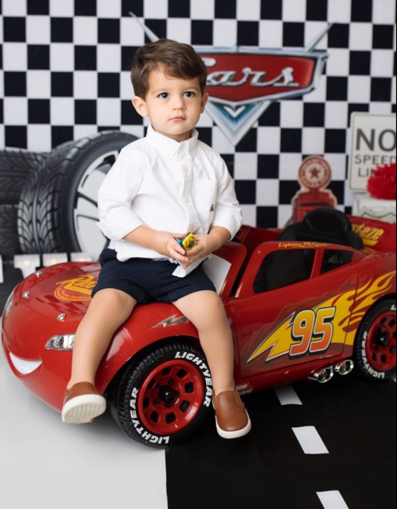 Child sitting on large Lightning McQueen toy car against checkered background with Cars movie theme decor.