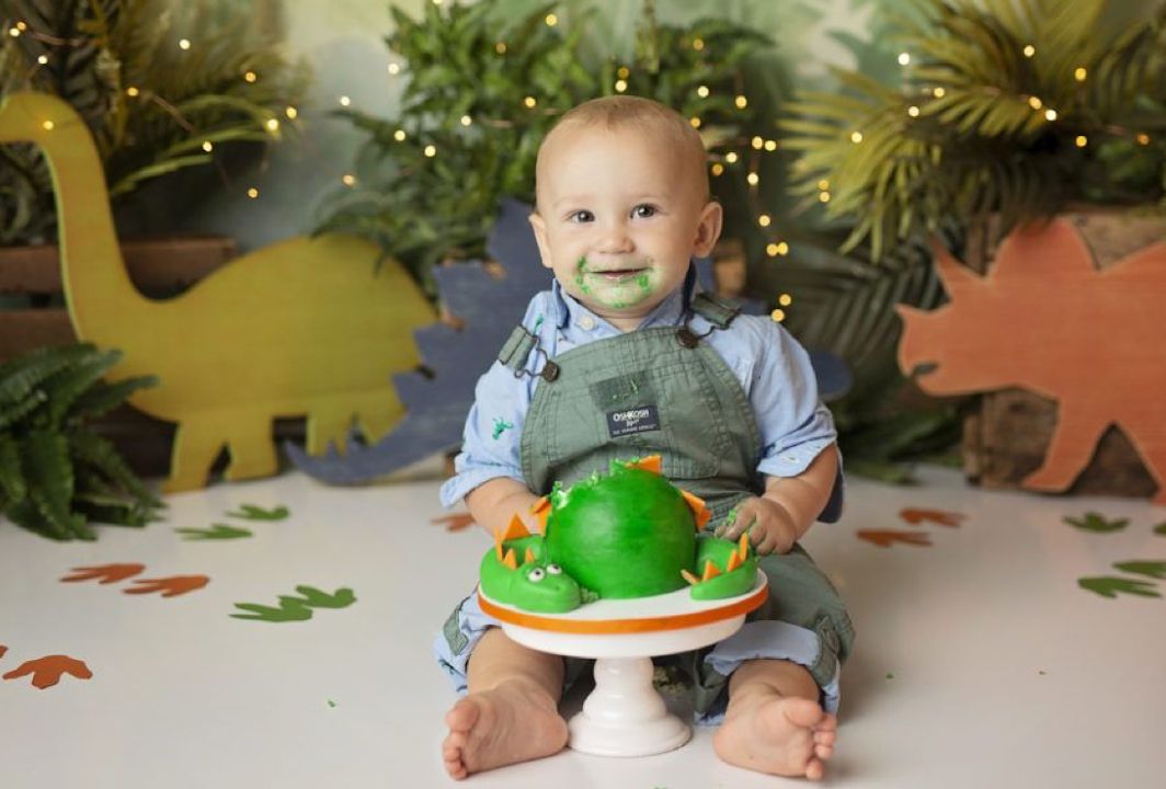 Baby in overalls sits with dinosaur decorations and green balloons at a first birthday photo session.