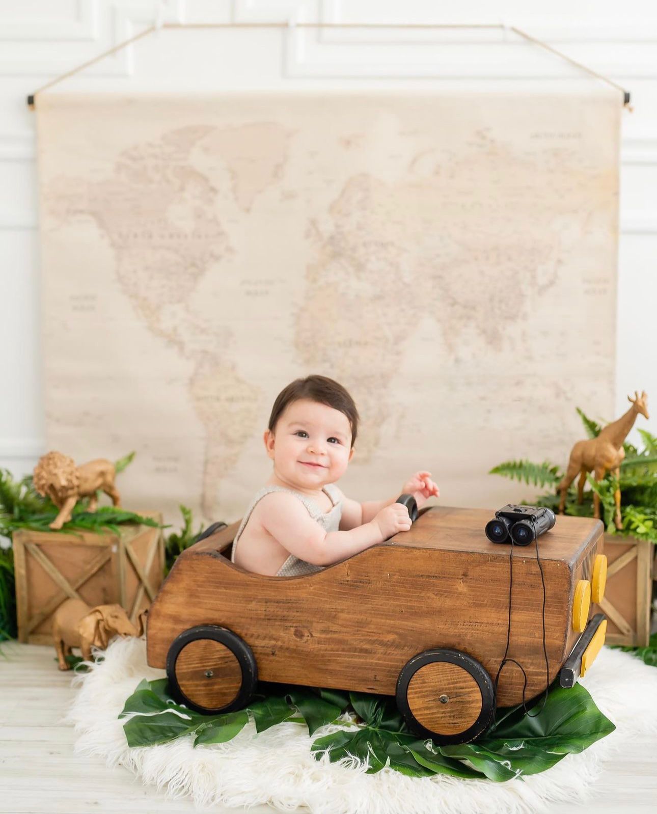 Baby in a wooden cart with greenery and toy animals in a vintage-themed photography setup against a world map backdrop.