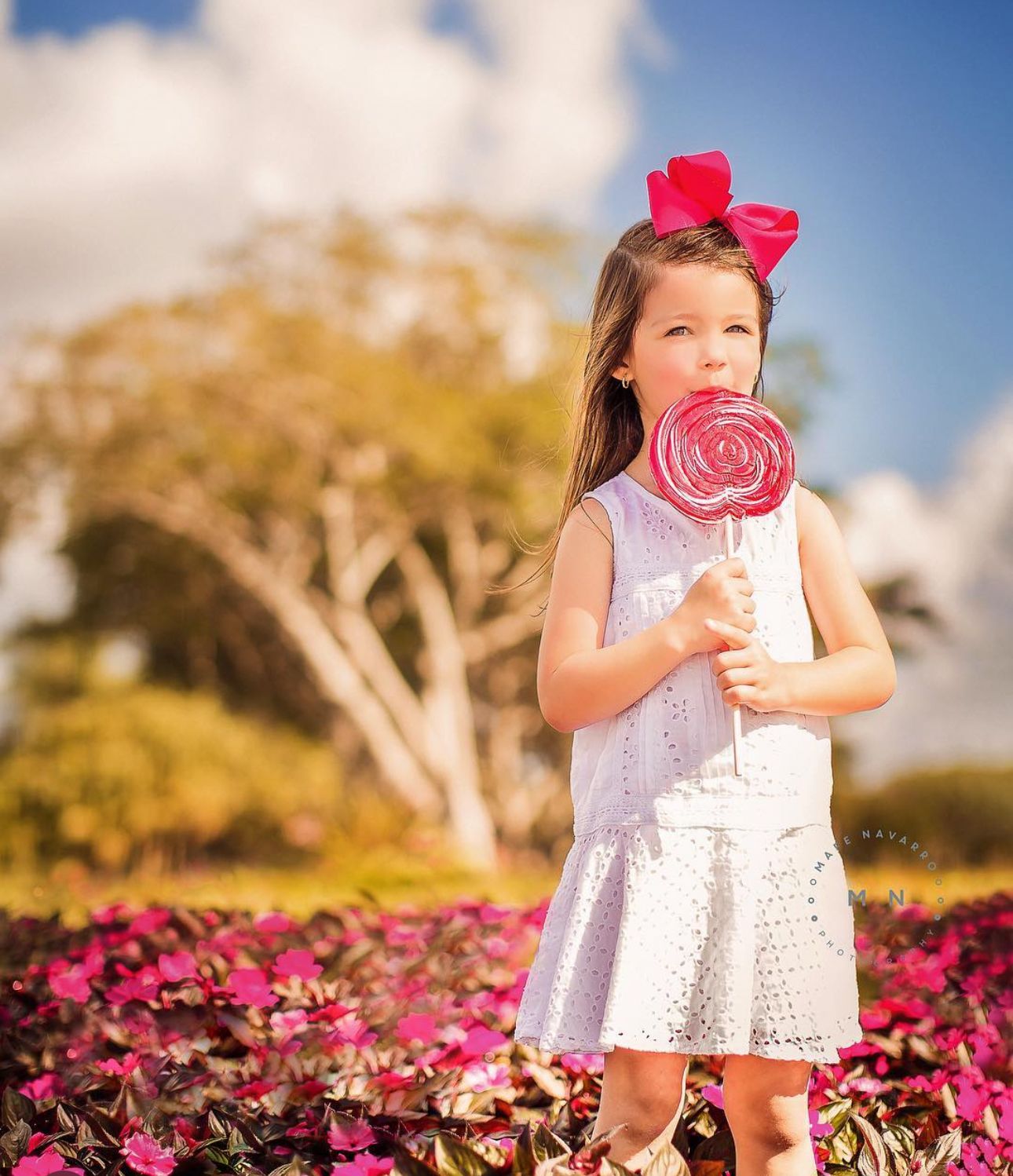 Young girl in white dress holding large pink lollipop, wearing pink bow, standing in colorful flower garden outdoors.