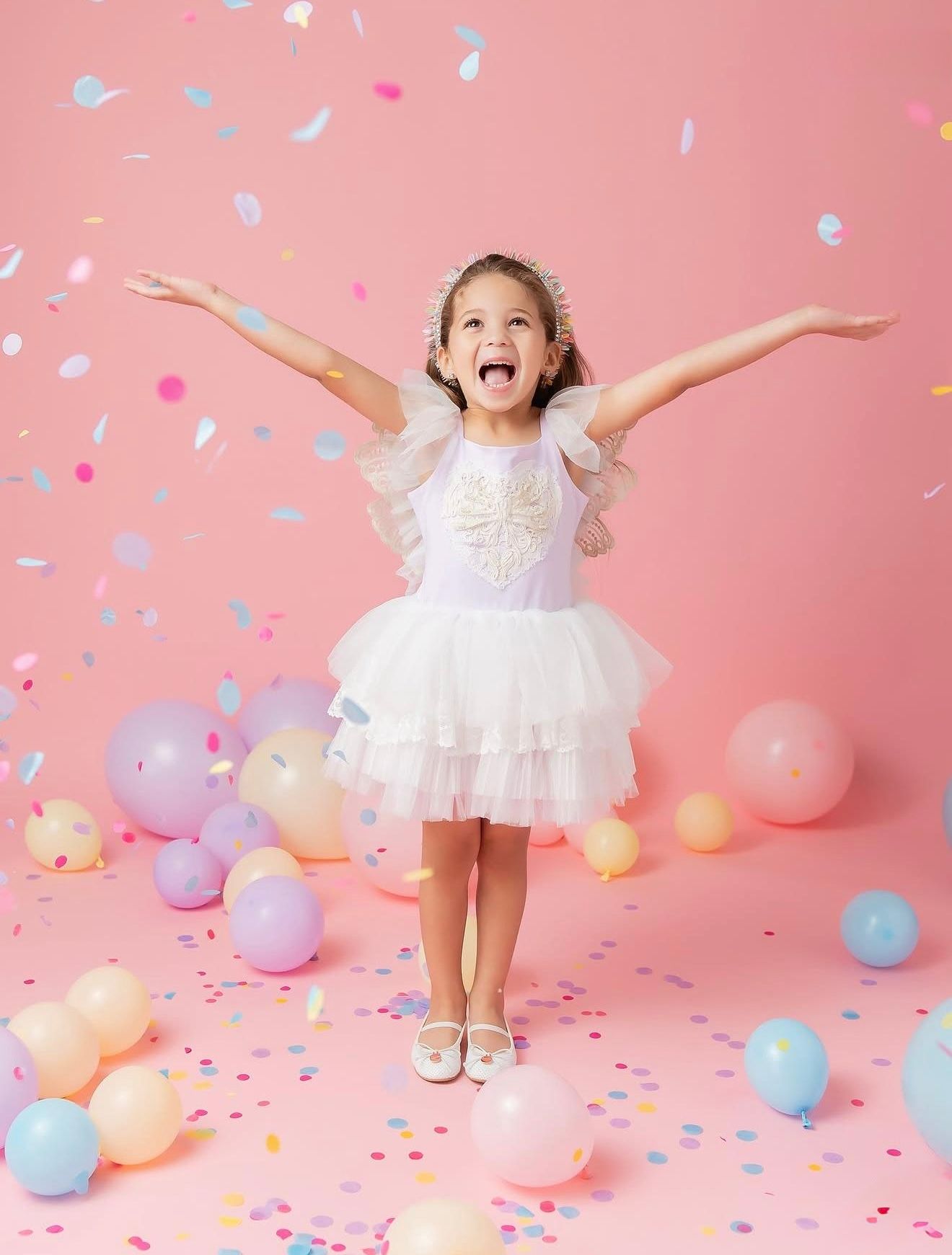 A joyful girl in a white tulle dress celebrates with confetti and pastel balloons on a pink background.