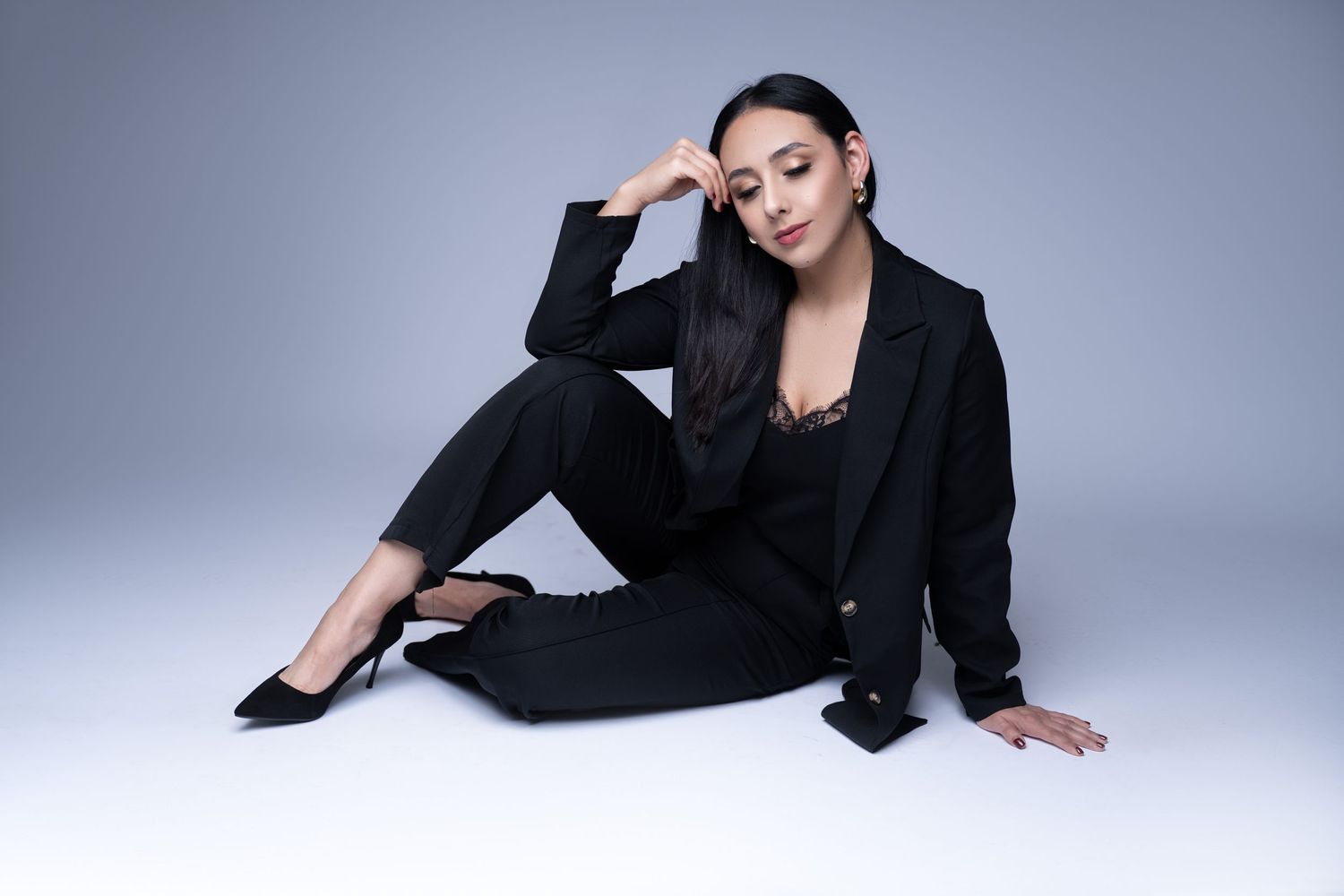 Fashion model in all black ensemble posing on floor against gray studio background.