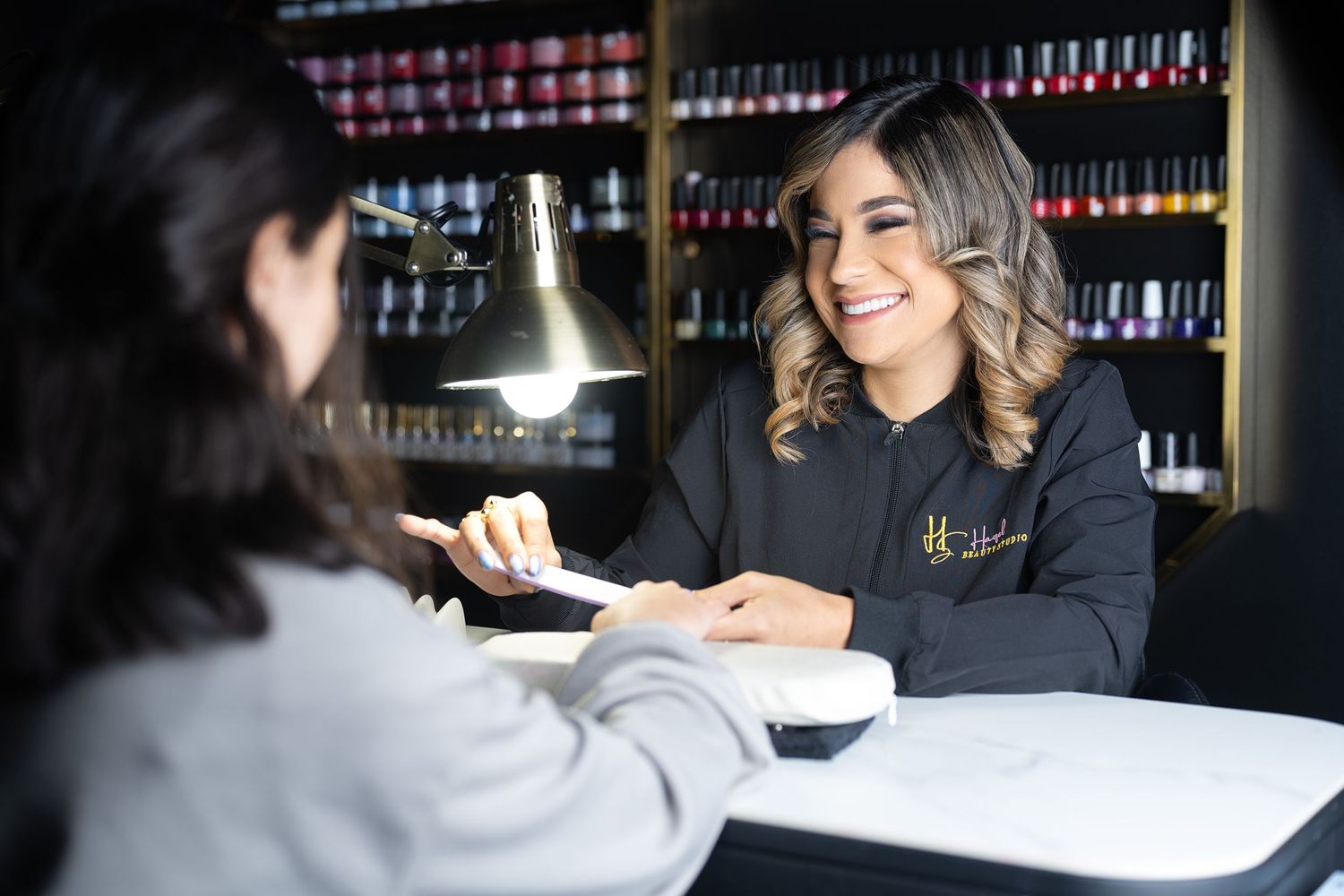 Professional nail technician consulting with client at salon workstation with polish display wall in background.