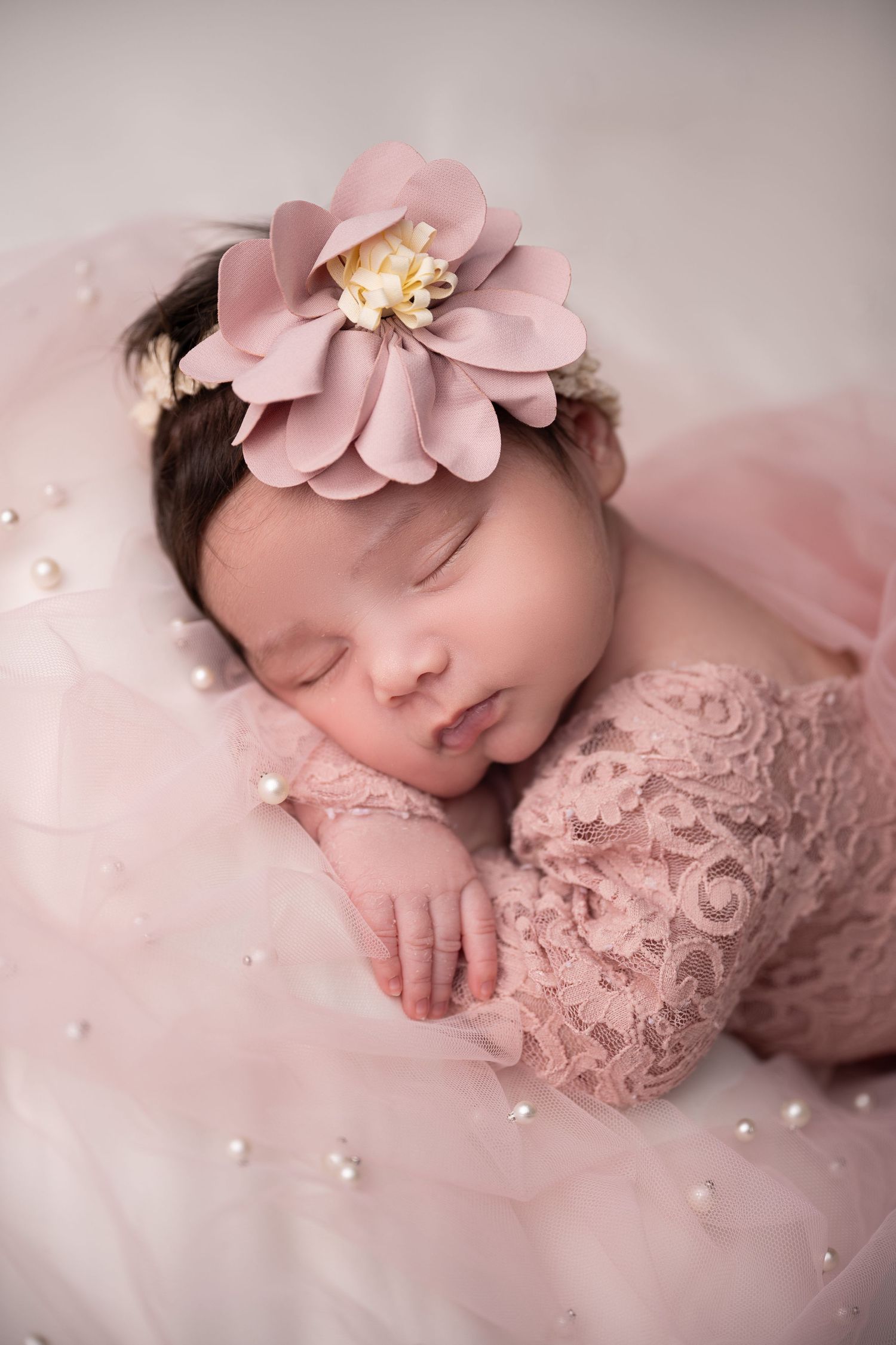 Sleeping newborn in pink lace wearing large floral headpiece against rosy backdrop.